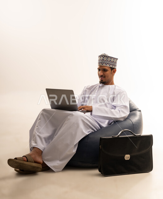 تصفح الانترنت وبرامج التواصل الاجتماعي, إستخدام الحاسوب المحمول لإنجاز المهام، بورتريه لرجل عربي خليجي عماني يرتدي الدشداشه والكمه يجلس على أريكه مريحه يعمل على جهاز الابتوب، تطور التكنولوجيا والأجهزة التقنية، خلفية بيضاء