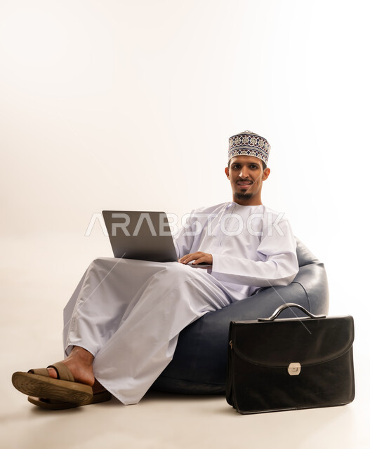 تصفح الانترنت وبرامج التواصل الاجتماعي, إستخدام الحاسوب المحمول لإنجاز المهام، بورتريه لرجل عربي خليجي عماني يرتدي الدشداشه والكمه يجلس على أريكه مريحه يضع جهاز الابتوب على ساقيه ينظر ناحيه شئ ما، تطور التكنولوجيا والأجهزة التقنية، خلفية بيضاء