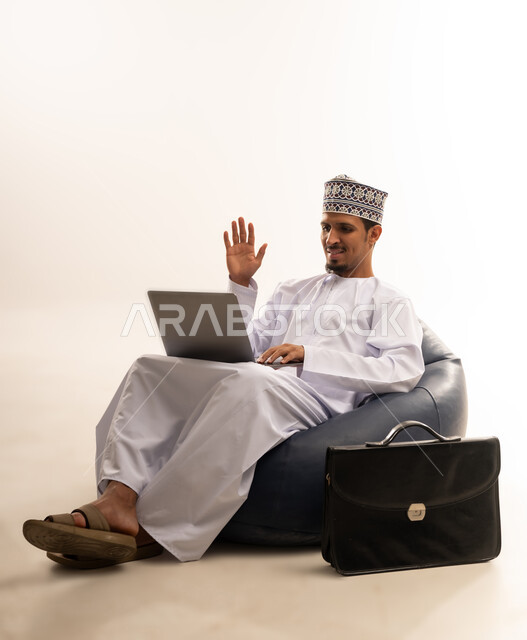 تصفح الانترنت وبرامج التواصل الاجتماعي, إستخدام الحاسوب المحمول لإنجاز المهام، بورتريه لرجل عربي خليجي عماني يرتدي الدشداشه والكمه يجلس على أريكه مريحه  يشير بعلامه التوقف عن فعل شئ ما أمام شاشه الابتوب, تطور التكنولوجيا والأجهزة التقنية، خلفية بيضاء