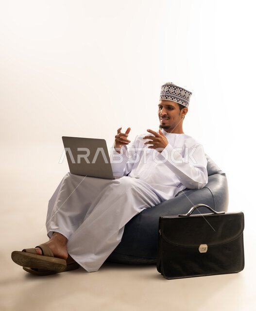 تصفح الانترنت وبرامج التواصل الاجتماعي, إستخدام الحاسوب المحمول لإنجاز المهام، بورتريه لرجل عربي خليجي عماني يرتدي الدشداشه والكمه يجلس على أريكه مريحه  يشير بيديه بعلامات التعجب من شئ ما بشاشه الابتوب, تطور التكنولوجيا والأجهزة التقنية، خلفية بيضاء