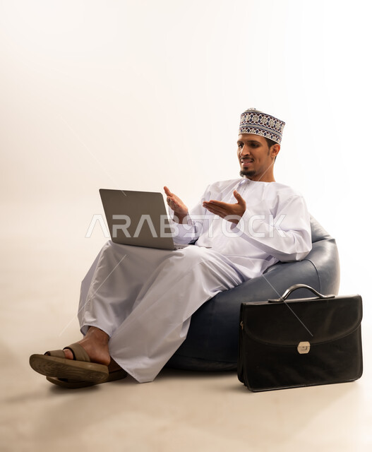 Browsing the internet and social networking programs, using a laptop to complete tasks, a portrait of an Arab Gulf Omani man wearing a dishdasha and a kuma sitting on a comfortable sofa, pointing his hands towards the laptop screen with a smile, talking about something, the development of technology and technical devices, white background