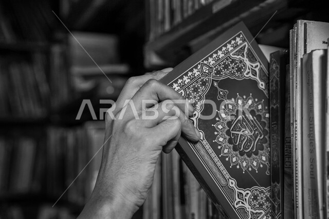 قراءة وتلاوة كلام الله عز وجل من المصحف الشريف، صورة مقربة بالأبيض والاسود ليد رجل عربي خليجي سعودي يقوم بأخذ القرآن الكريم من احدى رفوف المكتبة، العبادة والتقرب الى الله
