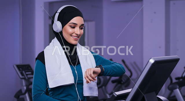 التدرب بعزيمة للمحافظة على نظام الحياة الصحي، ايماءات السعادة والنشاط، مفهوم الرشاقة واللياقة، صورة مقربة لفتاة عربية خليجية سعودية ترتدي الملابس الرياضية تضع الهيدفون على رأسها تتتابع تقدمها في الأداء على جهاز المشي، ممارسة الهوايات المفيدة