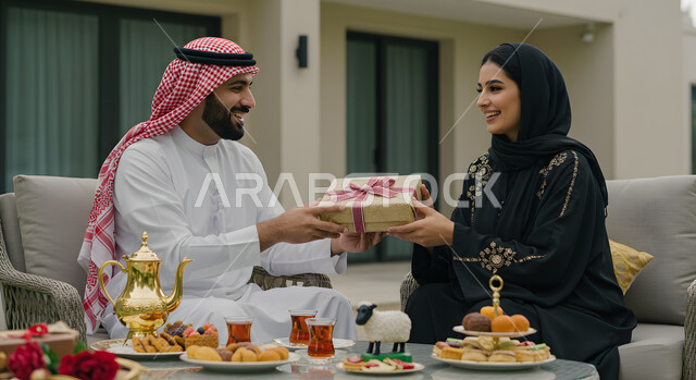 إحياء ذكرى يوم الزواج, جلسات ممتعه خارج المنزل, رجل عربي خليجي سعودي يرتدي الشماغ والثوب التقليدي يقدم لزوجته هديه في علبه مغلفة بمناسبة عيد الحب, الجلوس في الهواء الطلق في إحدى المنتجعات الفاخره, التعبير عن الألفة والمحبة بين الزوجين, إيماءات السعادة والسرور