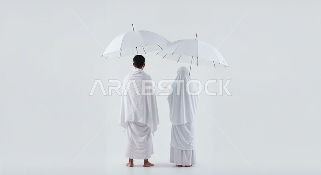 التظلل من اشعة الشمس باستخدام المظلة، مفهوم أركان الإسلام وأداء الفرائض الدينية، بورتريه من الخلف لحاج خليجي عربي سعودي يرتدي ثياب الإحرام يقف بجانب زوجته باستقامة بتعابير السرور، تأدية مناسك الحج والعمرة، العبادة والتقرب إلى الله، الجسم بالطول الكامل، خلفية بيضاء