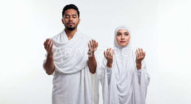 رفع اليدين والدعاء بخشوع وتضرع، مفهوم أركان الإسلام وأداء الفرائض الدينية، بورتريه مقرب لشاب حاج خليجي عربي سعودي يرتدي ثياب الإحرام يقف بجانب زوجته باستقامة بتعابير السرور، تأدية مناسك الحج والعمرة، العبادة والتقرب إلى الله، خلفية بيضاء