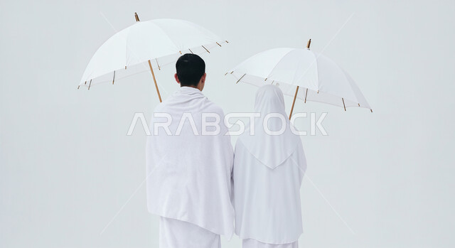 التظلل من اشعة الشمس باستخدام المظلة، مفهوم أركان الإسلام وأداء الفرائض الدينية، بورتريه مقرب من الخلف لشاب حاج خليجي عربي سعودي يرتدي ثياب الإحرام يقف بجانب زوجته باستقامة بتعابير السرور، تأدية مناسك الحج والعمرة، العبادة والتقرب إلى الله، خلفية بيضاء