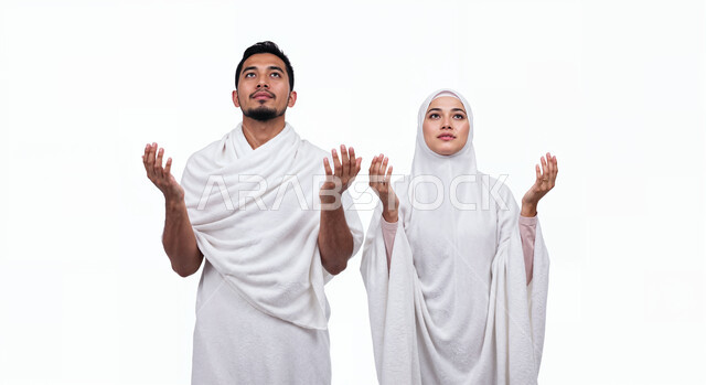 رفع اليدين والدعاء بخشوع وتضرع، مفهوم أركان الإسلام وأداء الفرائض الدينية، بورتريه مقرب لشاب حاج خليجي عربي سعودي يرتدي ثياب الإحرام يقف بجانب زوجته باستقامة بتعابير السرور، تأدية مناسك الحج والعمرة، العبادة والتقرب إلى الله، خلفية بيضاء