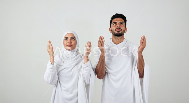 رفع اليدين والدعاء بخشوع وتضرع، مفهوم أركان الإسلام وأداء الفرائض الدينية، بورتريه مقرب لشاب حاج خليجي عربي سعودي يرتدي ثياب الإحرام يقف بجانب زوجته باستقامة بتعابير السرور، تأدية مناسك الحج والعمرة، العبادة والتقرب إلى الله، خلفية بيضاء