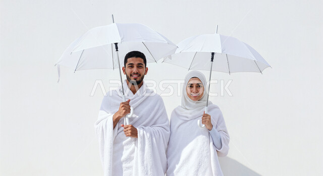 التظلل من اشعة الشمس باستخدام المظلة، مفهوم أركان الإسلام وأداء الفرائض الدينية، بورتريه مقرب لشاب حاج خليجي عربي سعودي يرتدي ثياب الإحرام يقف بجانب زوجته باستقامة بتعابير السرور، تأدية مناسك الحج والعمرة، العبادة والتقرب إلى الله، خلفية بيضاء