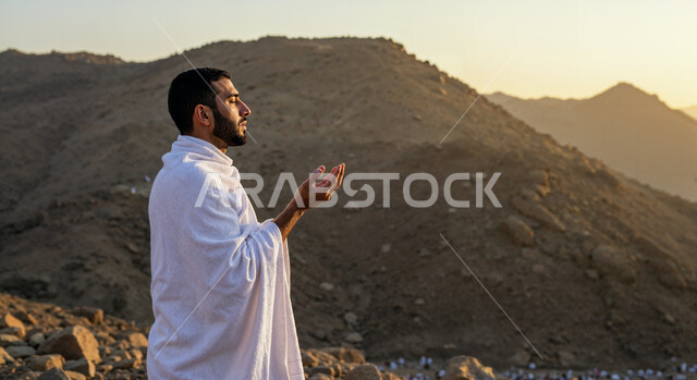 الوقوف على جبل عرفات، العبادة والتقرب إلى الله، مفهوم أركان الإسلام وأداء الفرائض الدينية، رفع اليدين والدعاء بخشوع وتضرع، صورة مقربة من الجانب لرجل حاج خليجي عربي سعودي يرتدي ثياب الإحرام يقوم بتأدية مناسك الحج والعمرة