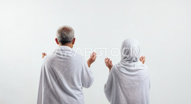 رفع اليدين والدعاء بخشوع وتضرع، مفهوم أركان الإسلام وأداء الفرائض الدينية، بورتريه مقرب من الخلف لحاج كبير بالسن خليجي عربي سعودي يرتدي ثياب الإحرام يقف بجانب زوجته باستقامة، تأدية مناسك الحج والعمرة، العبادة والتقرب إلى الله، خلفية بيضاء