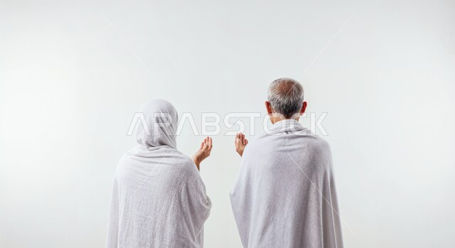 رفع اليدين والدعاء بخشوع وتضرع، مفهوم أركان الإسلام وأداء الفرائض الدينية، بورتريه مقرب من الخلف لحاج كبير بالسن خليجي عربي سعودي يرتدي ثياب الإحرام يقف بجانب زوجته باستقامة، تأدية مناسك الحج والعمرة، العبادة والتقرب إلى الله، خلفية بيضاء