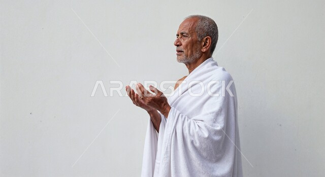 مفهوم أركان الإسلام وأداء الفرائض الدينية، رفع اليدين والدعاء بخشوع وتضرع، بورتريه مقرب من الجانب لحاج كبير بالسن خليجي عربي سعودي يرتدي ثياب الإحرام يقوم بتأدية مناسك الحج والعمرة، العبادة والتقرب إلى الله، خلفية بيضاء
