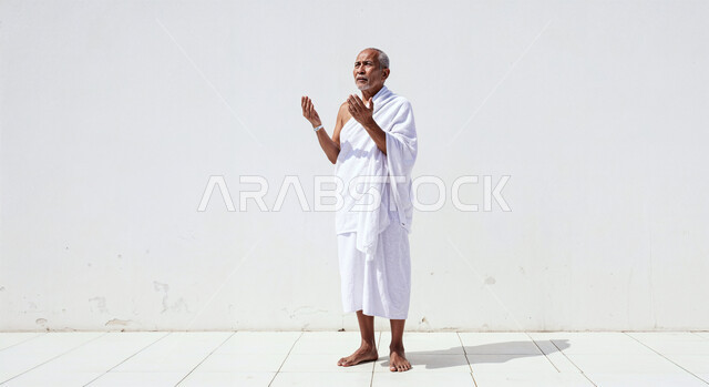 مفهوم أركان الإسلام وأداء الفرائض الدينية، رفع اليدين والدعاء بخشوع وتضرع، بورتريه لحاج كبير بالسن خليجي عربي سعودي يرتدي ثياب الإحرام يقوم بتأدية مناسك الحج والعمرة، العبادة والتقرب إلى الله، صورة الجسم بالطول الكامل، خلفية بيضاء