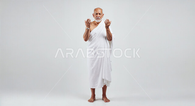 مفهوم أركان الإسلام وأداء الفرائض الدينية، رفع اليدين والدعاء بخشوع وتضرع، بورتريه لحاج كبير بالسن خليجي عربي سعودي يرتدي ثياب الإحرام يقوم بتأدية مناسك الحج والعمرة، العبادة والتقرب إلى الله، صورة الجسم بالطول الكامل، خلفية بيضاء