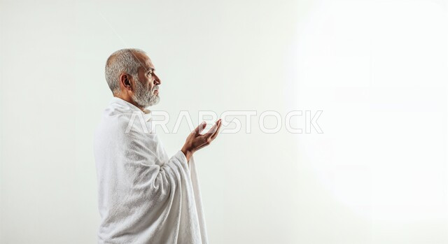 مفهوم أركان الإسلام وأداء الفرائض الدينية، رفع اليدين والدعاء بخشوع وتضرع، بورتريه مقرب من الجانب لحاج كبير بالسن خليجي عربي سعودي يرتدي ثياب الإحرام يقوم بتأدية مناسك الحج والعمرة، العبادة والتقرب إلى الله، خلفية بيضاء