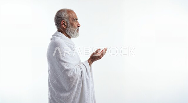 مفهوم أركان الإسلام وأداء الفرائض الدينية، رفع اليدين والدعاء بخشوع وتضرع، بورتريه مقرب من الجانب لحاج كبير بالسن خليجي عربي سعودي يرتدي ثياب الإحرام يقوم بتأدية مناسك الحج والعمرة، العبادة والتقرب إلى الله، خلفية بيضاء
