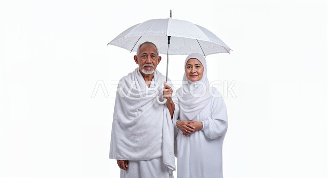 Shading from the sun using an umbrella, the concept of the pillars of Islam and performing religious duties, close-up portrait of an elderly Saudi Arabian Gulf pilgrim wearing Ihram clothes standing beside his wife straight with happy expressions, performing Hajj and Umrah rituals, worship and getting closer to God, white background