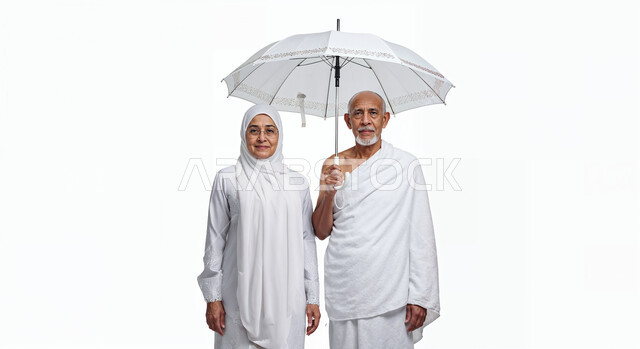 التظلل من اشعة الشمس باستخدام المظلة، مفهوم أركان الإسلام وأداء الفرائض الدينية، بورتريه مقرب لحاج كبير بالسن خليجي عربي سعودي يرتدي ثياب الإحرام يقف بجانب زوجته باستقامة بتعابير السرور، تأدية مناسك الحج والعمرة، العبادة والتقرب إلى الله، خلفية بيضاء