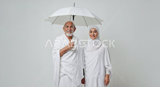 التظلل من اشعة الشمس باستخدام المظلة، مفهوم أركان الإسلام وأداء الفرائض الدينية، بورتريه مقرب لحاج كبير بالسن خليجي عربي سعودي يرتدي ثياب الإحرام يقف بجانب زوجته باستقامة بتعابير السرور، تأدية مناسك الحج والعمرة، العبادة والتقرب إلى الله، خلفية بيضاء