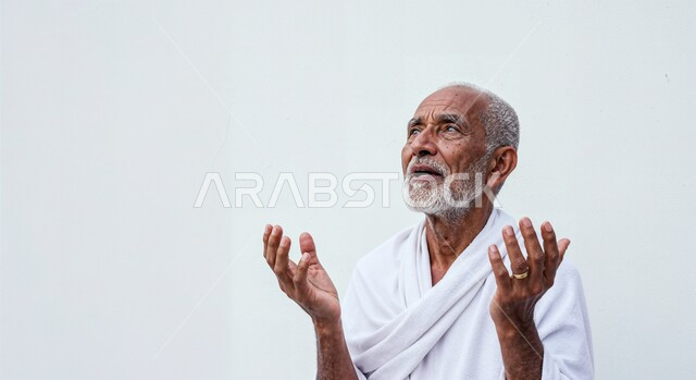مفهوم أركان الإسلام وأداء الفرائض الدينية، رفع اليدين والدعاء بخشوع وتضرع، بورتريه مقرب لحاج كبير بالسن خليجي عربي سعودي يرتدي ثياب الإحرام يقوم بتأدية مناسك الحج والعمرة، العبادة والتقرب إلى الله، خلفية بيضاء