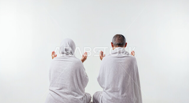 رفع اليدين والدعاء بخشوع وتضرع، مفهوم أركان الإسلام وأداء الفرائض الدينية، بورتريه مقرب من الخلف لحاج كبير بالسن خليجي عربي سعودي يرتدي ثياب الإحرام يقف بجانب زوجته باستقامة، تأدية مناسك الحج والعمرة، العبادة والتقرب إلى الله، خلفية بيضاء