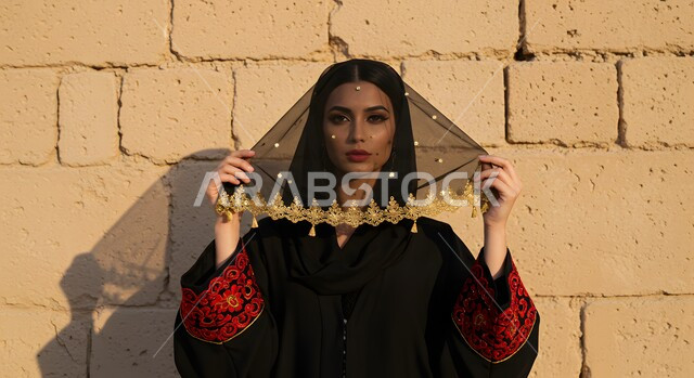 يوم بدينا 1727 م, ذكرى تأسيس الدولة السعودية الأولى في 22 فبراير, التباهي بإنجازات الوطن, صورة  مقربة لشابة عربية خليجية سعودية ترتدي المقطع والهامة تنظر إلى الكاميرا بتعابير الثقة بالنفس, ارتداء زي المنطقة الوسطى في المناسبات الوطنية, الوقوف امام بيت تراثي شعبي طيني
