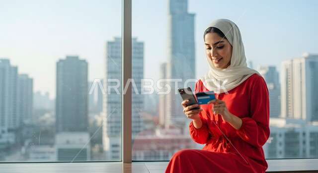 Shopping and paying through mobile programs and applications, the ability to pay bills via credit card, a smiling Saudi Arabian Gulf woman wearing the hijab holding a mobile phone and a bank credit card, lifestyle and quality of life in the Kingdom, expressions of pleasure using modern technologies and means