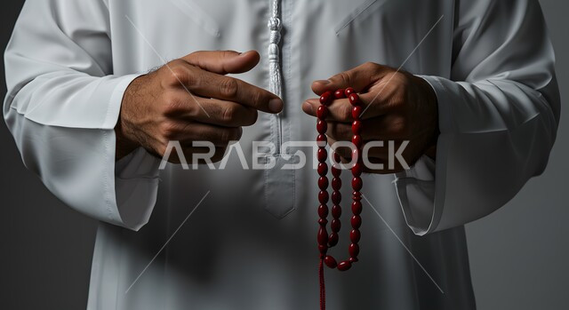  العبادة والطاعة والتقرب الى الله، بورتريه مقرب ليد رجل عربي خليجي اماراتي يرتدي الكندورة يمسك بيده سبحة من حبات من الخرز الحمراء، التسبيح والتهليل وذكر الله تعالى، الاجتهاد في العبادات, خلفية رمادية
