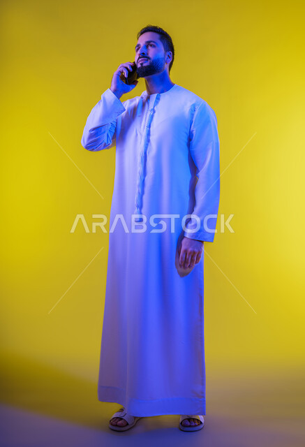 اجراء مكالمة هاتفية من خلال الجوال، بورتريه لرجل عربي خليجي إماراتي يرتدي الكندورة الإماراتية ، استخدام الهاتف المحمول في الاتصال والتواصل مع الأفراد، دردشة ومحادثات وتعارف، خلفية صفراء .