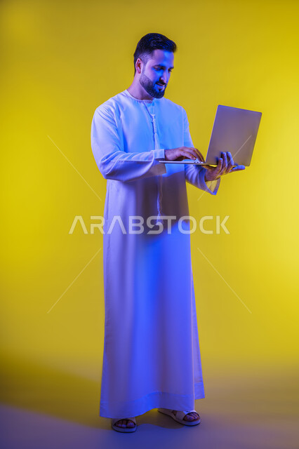 دمج العمل مع التقنيات والتكنولوجيا ، إدارة شؤون الأعمال وتنظيمها عبر الأجهزة التقنية الحديثة، مفهوم تسهيل الاعمال والقدرة على إنجاز المهام عن بعد، بورتريه لشاب عربي خليجي اماراتى يرتدي الكندورة يمسك بيده حاسوب محمول تظهر عليه علامات الفرح والسرور، الجسم بالطول الكامل، خلفية صفراء .