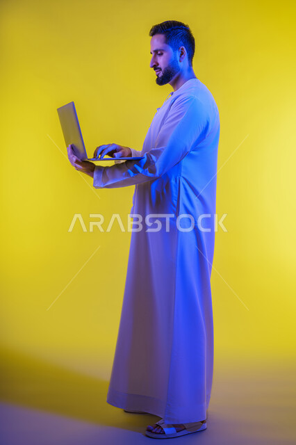 إدارة شؤون الأعمال وتنظيمها عبر الأجهزة التقنية الحديثة، مفهوم تسهيل الاعمال والقدرة على إنجاز المهام عن بعد، بورتريه من الجانب لشاب عربي خليجي اماراتى يرتدي الكندورة يمسك بيده حاسوب محمول تظهر عليه علامات الفرح والسرور، الجسم بالطول الكامل، خلفية صفراء .