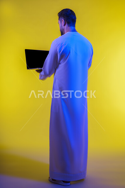 Using modern technologies and advanced devices, the concept of facilitating work and the ability to complete tasks remotely, a portrait from the back of an Arab Gulf Emirati young man wearing a kandura holding a laptop, showing signs of joy and happiness, full body, yellow background.