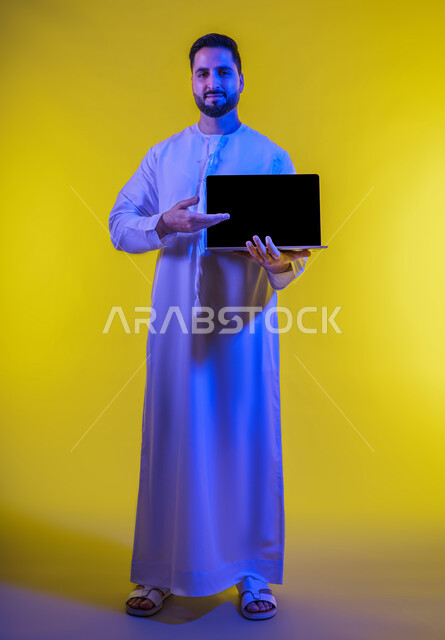 مفهوم تسهيل الاعمال والقدرة على إنجاز المهام عن بعد، استخدام التقنيات الحديثة والأجهزة المتطورة ، بورتريه لشاب عربي خليجي اماراتى يرتدي الكندورة يمسك بيده حاسوب محمول تظهر عليه علامات الفرح والسرور، الجسم بالطول الكامل، خلفية صفراء .