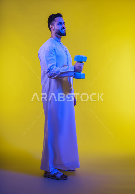 رفع الاوزان وعرض عضلات اليد ، التمتع بالبنية الجسدية القوية، بورتريه لرجل عربي خليجي إماراتي يرتدي الكندورة الإماراتية ، إيماءات وجه ويدين تدل على القوة العضلية، اللياقة البدنية والجسم الصحي المتين، خلفية صفراء .