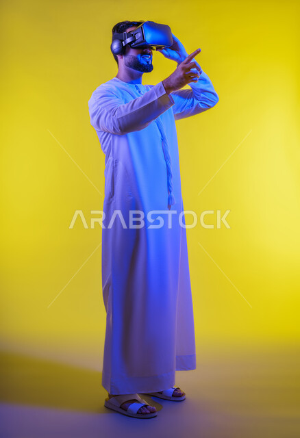الاستمتاع في قضاء الأوقات الترفيهية عبر استخدام التقنيات الحديثة الالكترونية، دمج التكنولوجيا بالحياة العملية، الاندماج والتفاعل مع العالم الافتراضي،  بورتريه لشاب عربي خليجي إماراتي يرتدي الكندورة يضع نظارات الفي آر ثلاثية الابعاد، خلفية صفراء .