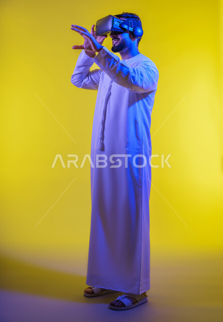 Integrating technology into practical life, merging and interacting with the virtual world, enjoying leisure time using modern electronic technologies, portrait of an Arab Gulf Emirati young man wearing a kandura and 3D VR glasses, yellow background.
