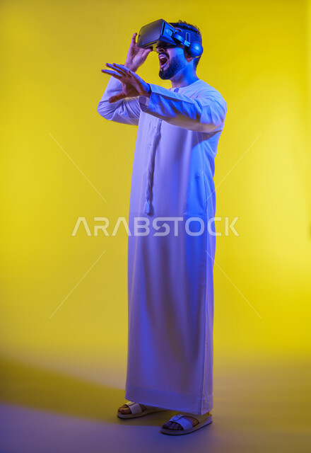 الاندماج والتفاعل مع العالم الافتراضي، دمج التكنولوجيا بالحياة العملية، الاستمتاع في قضاء الأوقات الترفيهية عبر استخدام التقنيات الحديثة الالكترونية، بورتريه لشاب عربي خليجي إماراتي يرتدي الكندورة يضع نظارات الفي آر ثلاثية الابعاد، خلفية صفراء .