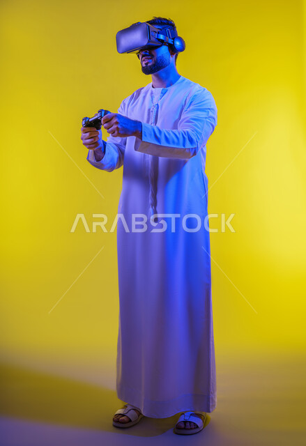 دمج العالم الخيالي مع المتعة والتسلية، بورتريه لرجل عربي خليجي اماراتي مبتسم يرتدي الكندورة التقليدية ونظارة الواقع الإفتراضي يمسك بيده عصا التحكم يقوم باللعب في ألعاب الفيديو الترفيهية، أجهزة تكنولوجية وتقنيات حديثة، صورة الجسم بالطول الكامل، خلفية صفراء .