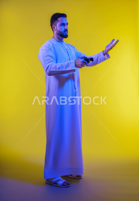 المتعة والتسلية فى العطله ، العاب الفيديو الترفيهية ، ايماءات الخسارة والهزيمة  ، بورتريه من الجانب لرجل عربي خليجي اماراتي يرتدي الكندورة يقف ويمسك بيده عصا التحكم بتعابير السعادة ، الجسم بالطول الكامل، خلفية صفراء .