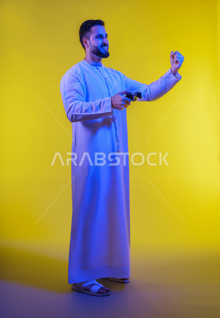 تقنيات والعاب حديثة ، قضاء وقت ممتع فى اوقات الفراغ ، العاب الفيديو الترفيهية ، ايماءات الخسارة والهزيمة  ، بورتريه من الجانب لرجل عربي خليجي اماراتي يرتدي الكندورة يقف ويمسك بيده عصا التحكم بتعابير السعادة ، الجسم بالطول الكامل، خلفية صفراء .