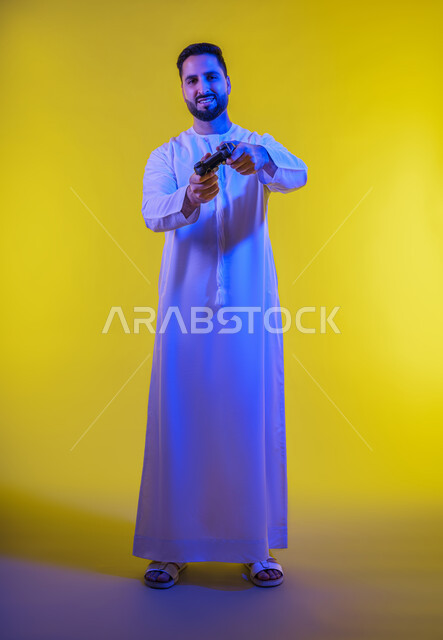 قضاء وقت ممتع فى اوقات الفراغ ، العاب الفيديو الترفيهية ، ايماءات الخسارة والهزيمة ،استخدام الأجهزة التكنولوجية والتقنيات الحديثة، ، بورتريه لرجل عربي خليجي اماراتي يرتدي الكندورة يقف ويمسك بيده عصا التحكم بتعابير السعادة ، الجسم بالطول الكامل، خلفية صفراء .