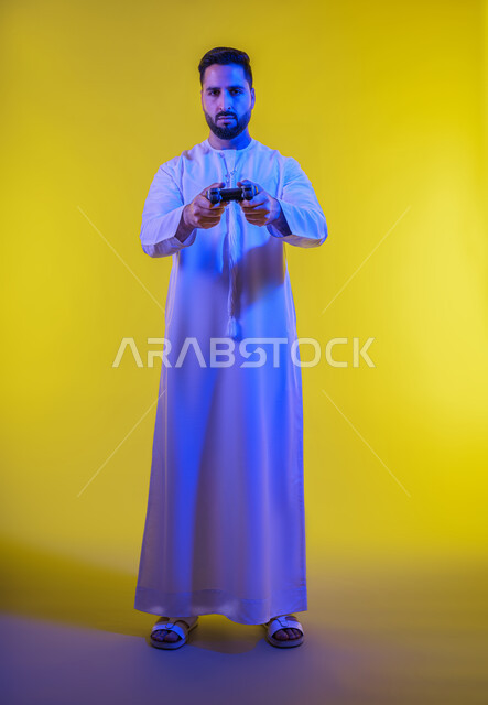 العاب الفيديو الترفيهية ، ايماءات الخسارة والهزيمة ،استخدام الأجهزة التكنولوجية والتقنيات الحديثة، ، بورتريه لرجل عربي خليجي اماراتي يرتدي الكندورة يقف ويمسك بيده عصا التحكم بتعابير السعادة ، الجسم بالطول الكامل، خلفية صفراء .