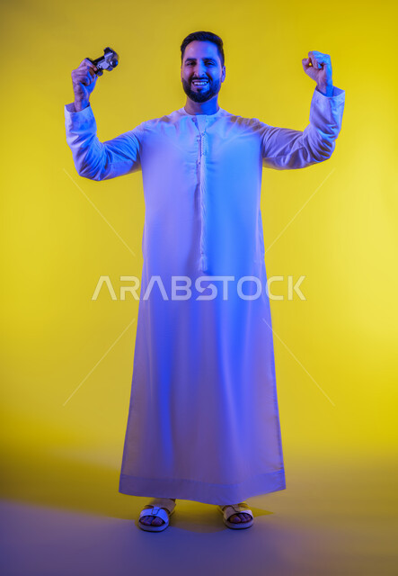 ايماءات الفوز والانتصار ، الاستمتاع باللعب بالعاب الفيديو الترفيهية، استخدام الأجهزة التكنولوجية والتقنيات الحديثة، بورتريه لرجل عربي خليجي اماراتي يرتدي الكندورة والغترة يقف ويمسك بيده عصا التحكم بتعابير السعادة ، الجسم بالطول الكامل، خلفية صفراء .