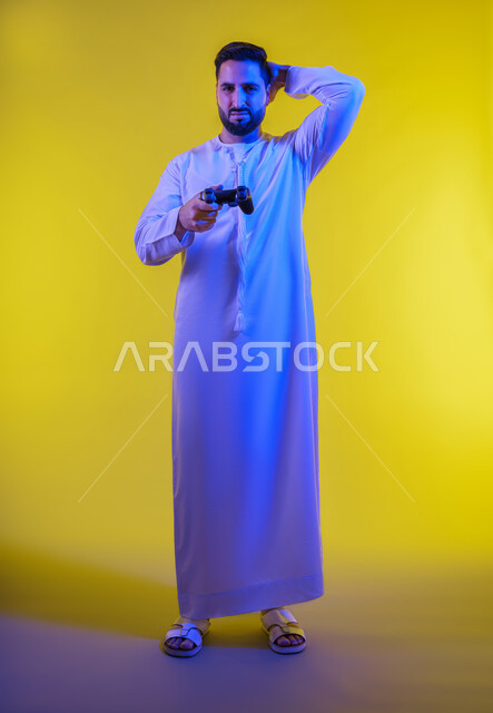 تعابير الاستمتاع باللعب بالألعاب الالكترونية الترفيهية، استخدام الأجهزة التكنولوجية والتقنيات الحديثة، بورتريه لرجل عربي خليجي اماراتي يرتدي الكندورة والغترة يقف ويمسك بيده عصا التحكم بإيماءات الخسارة ، الجسم بالطول الكامل، خلفية صفراء .