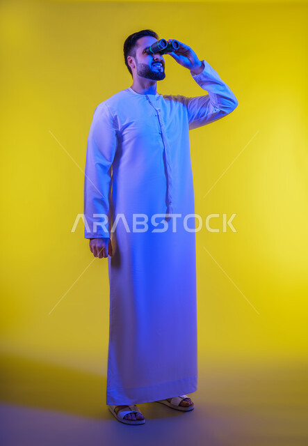 حب الاستكشاف و المغامرة, رؤية الاشياء البعيدة عبر التلسكوب, بورتريه لرجل عربي خليجي اماراتى مبتسم يرتدي الكندورة يحمل بيده منظار ثنائي العينين يشير بإصبع السبابة للأعلى ينظر إلى شيئ ما,  إيماءات الاستمتاع و السرور,  صورة الجسم بالطول الكامل, خلفية صفراء .