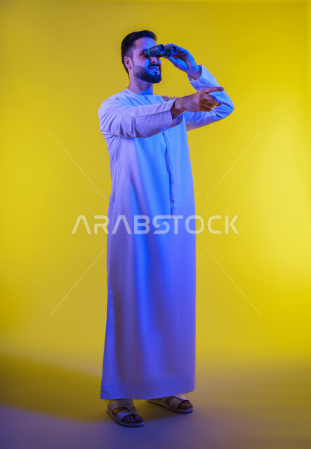 Seeing distant objects through a telescope, gestures of enjoyment and pleasure, portrait of a smiling Emirati Gulf Arab man wearing a kandura holding binoculars, pointing his index finger upwards looking at something, love of exploration and adventure, full body image, yellow background.