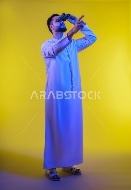 إيماءات الاستمتاع و السرور, حب الاستكشاف و المغامرة, بورتريه لرجل عربي خليجي اماراتى مبتسم يرتدي الكندورة يحمل بيده منظار ثنائي العينين يشير بإصبع السبابة للأعلى ينظر إلى شيئ ما, رؤية الاشياء البعيدة عبر التلسكوب, صورة الجسم بالطول الكامل, خلفية صفراء .