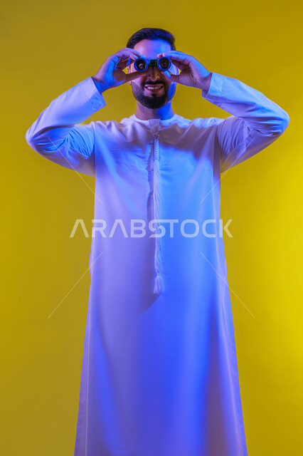 رؤية الاشياء البعيدة، حب المغامرة والاستكشاف، التخطيط المستقبلي للأهداف والطموحات، بورتريه لشاب عربي خليجي اماراتي يرتدي الكندورة يضع يده على عينه على شكل منظار، تعابير الفرح والسرور, خلفية صفراء .